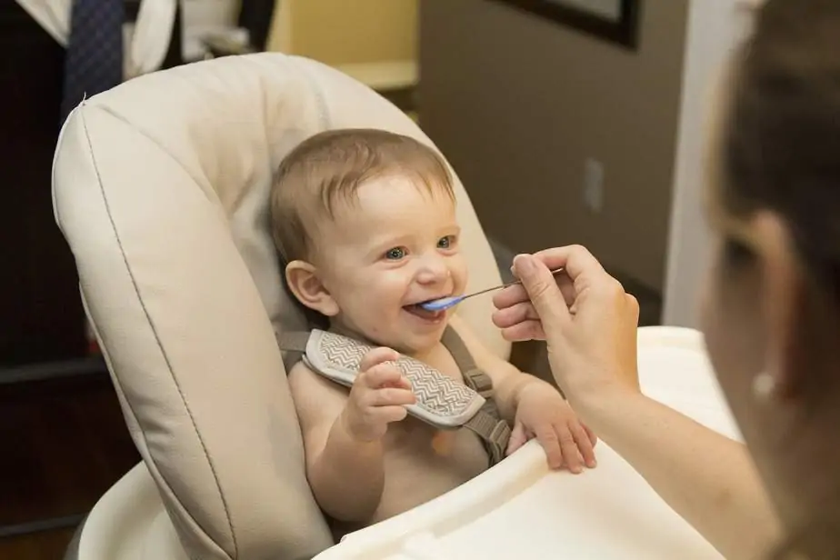 The baby boy seats in the kid chair and mommy feeds him with the spoon